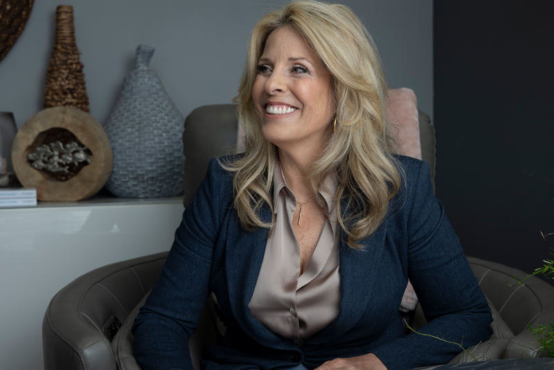 De'Anna Nuñez laughing in a charcoal blazer, seated in the armchair in her office