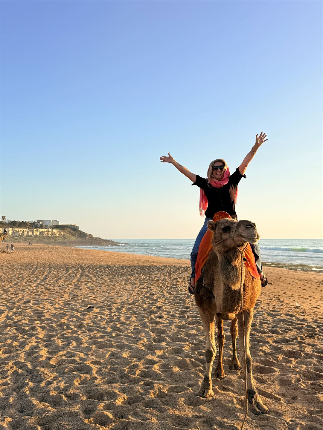 De'Anna Nuñez on a camel on a beach in Morocco, arms flung wide, laughing