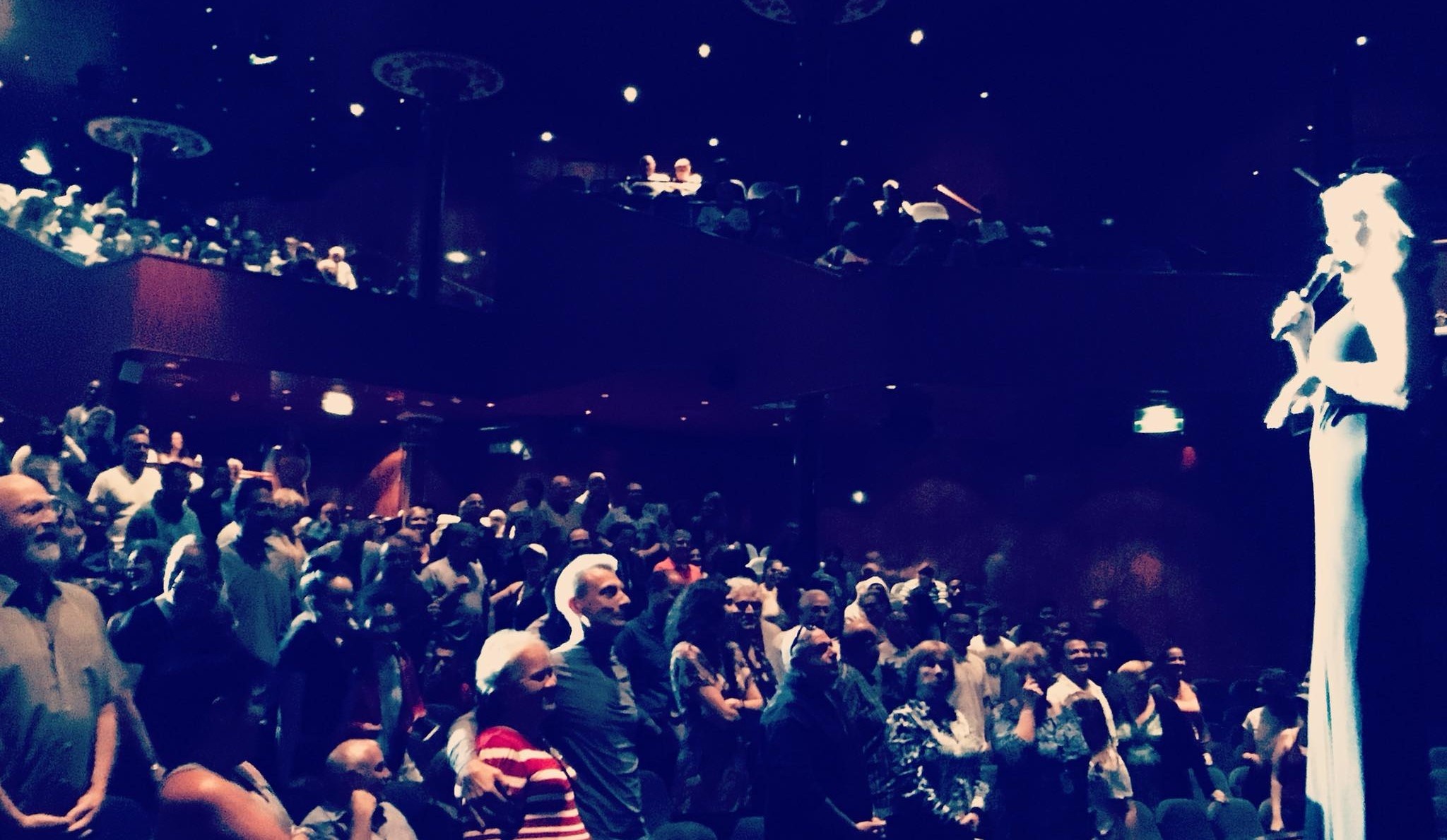 De'Anna Nuñez in a spotlight on stage, microphone in hand, speaking to a packed theater